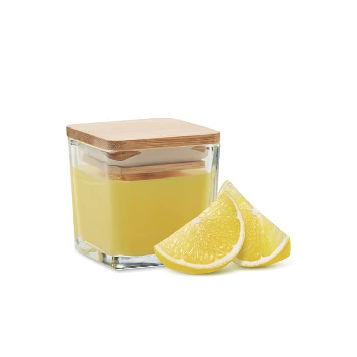 A yellow candle in a square glass container with a bamboo lid, accompanied by fresh lemon wedges.