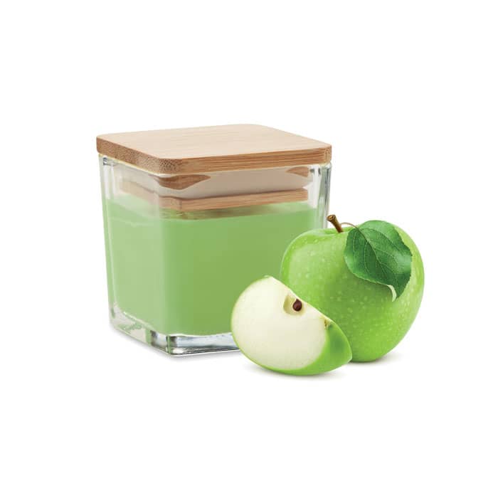 A green candle in a glass jar with a wooden lid, accompanied by a green apple and a slice of it.