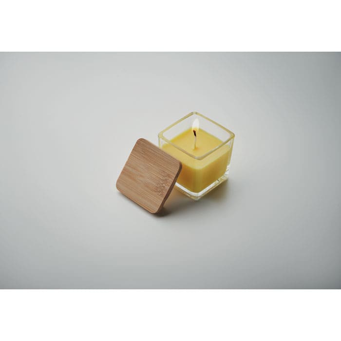 A yellow candle in a clear glass container with a wooden lid, positioned on a light grey background.