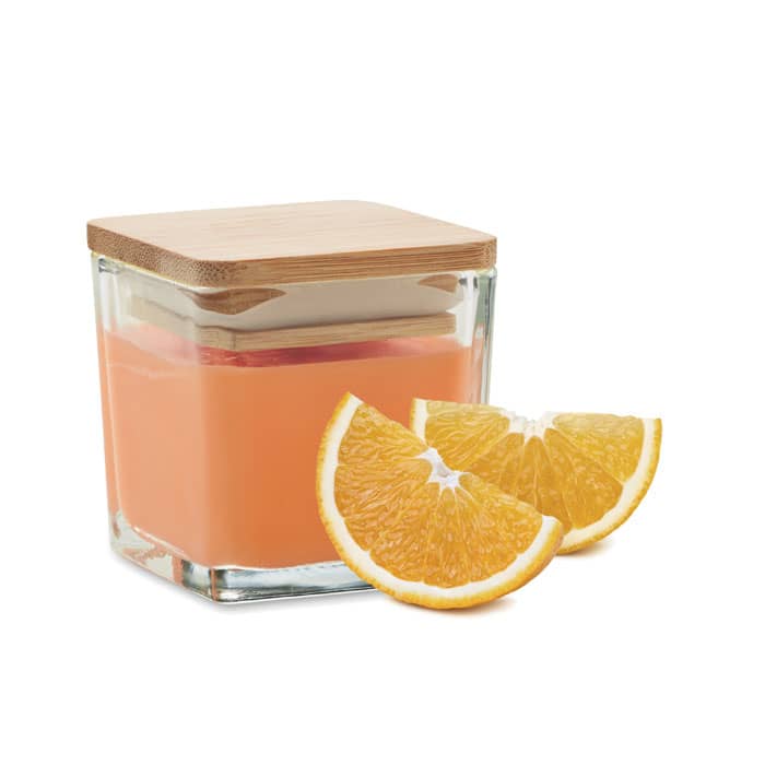 A glass jar candle with a bamboo lid, filled with orange wax, next to two orange slices.