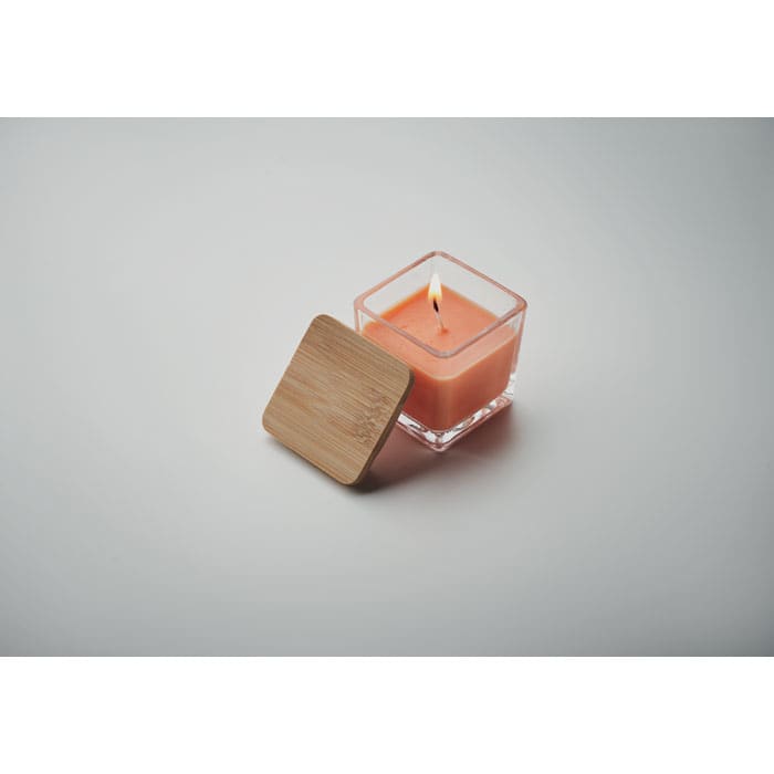 A candle in a square glass container with a light orange wax and a wooden lid, burning softly in the centre.
