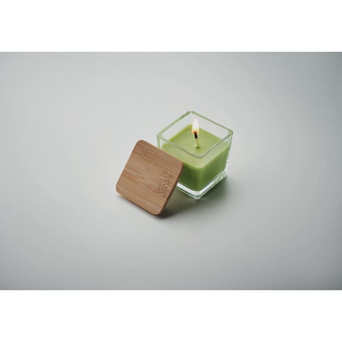 A green candle in a glass container with a bamboo lid, featuring a lit wick.