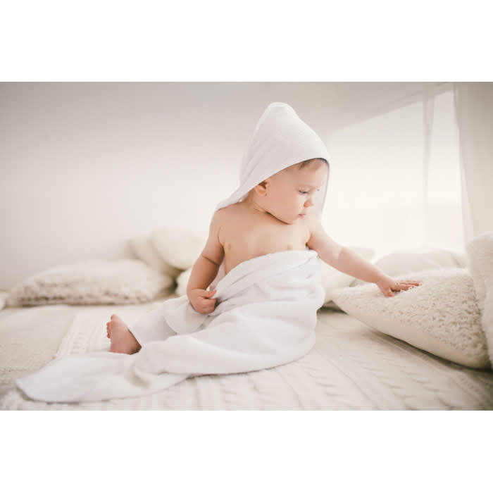 A baby wrapped in a white towel with a hood sits on a light-coloured bed surrounded by cushions.