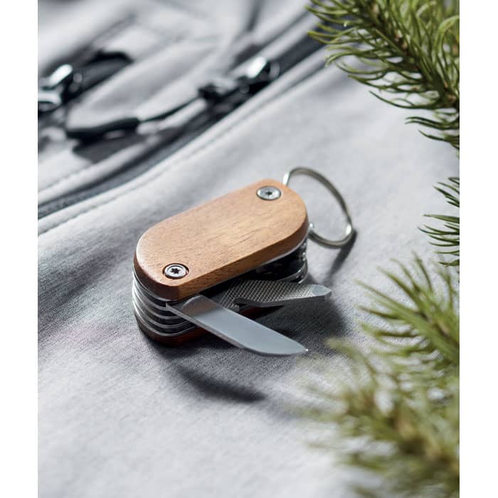 A multi-tool with a wooden handle, featuring two blades and a keyring, placed on a light grey surface alongside greenery.
