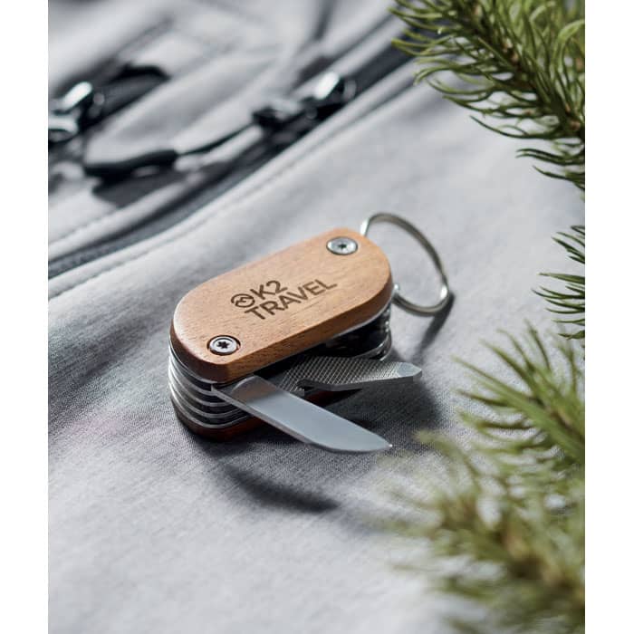 A multi-tool featuring a wooden handle and silver metal blades, resting on a grey fabric surface with greenery.