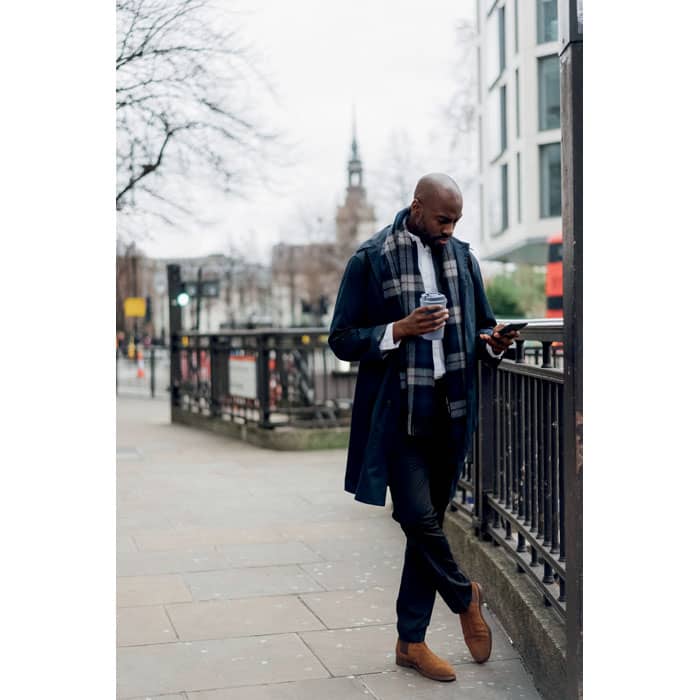 A man in a long navy coat and grey scarf holds a coffee cup while looking at his phone on a city street.