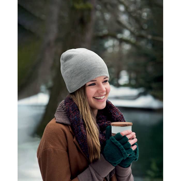 A woman wearing a grey beanie and a brown sweater holds a drink while smiling in a snowy, outdoor setting.