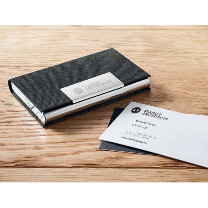 A black business card holder with a silver accent, displayed on a wooden surface next to a white business card.
