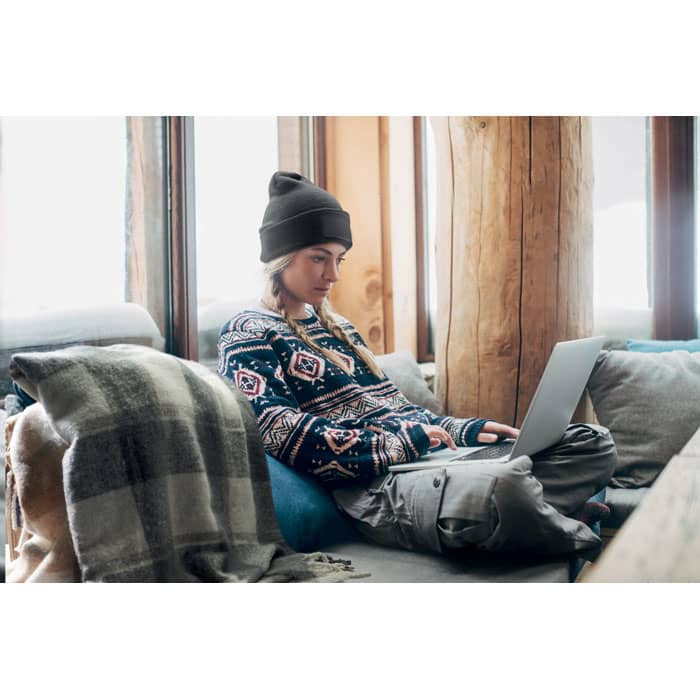 A young person in a patterned sweater and beanie sits on a couch, using a laptop near a window.