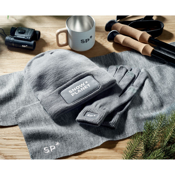 A grey beanie, gloves, and a scarf are laid out with a mug, binoculars, and ski poles. The items feature a logo.