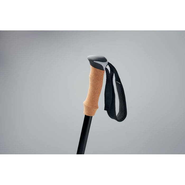A trekking pole with a black shaft, textured brown grip, and a wrist strap. The background is a soft grey.