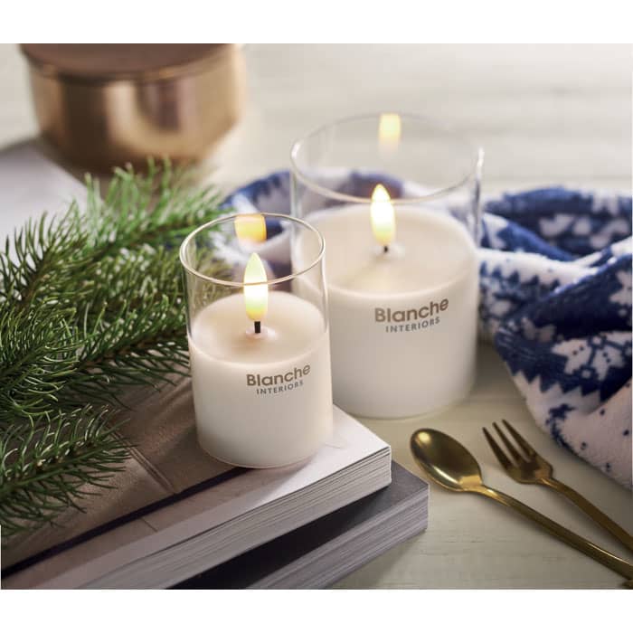 Two white candles in clear glass holders, with one candle lit, placed beside a blue patterned cloth and a small plant.