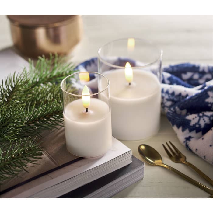 A set of two white candles in glass holders, surrounded by greenery, a patterned scarf, and decorative utensils.