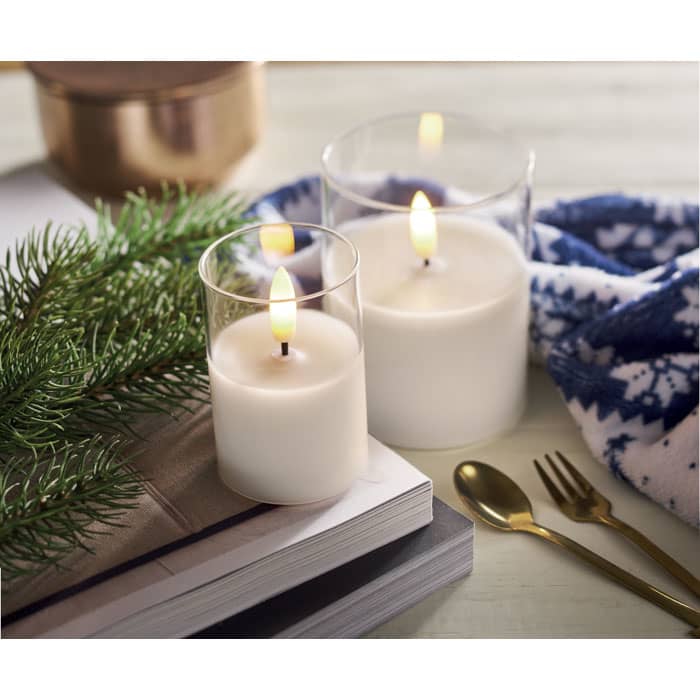 Two glass candles with flickering flames sit near greenery and a patterned fabric, alongside an open book and utensils.