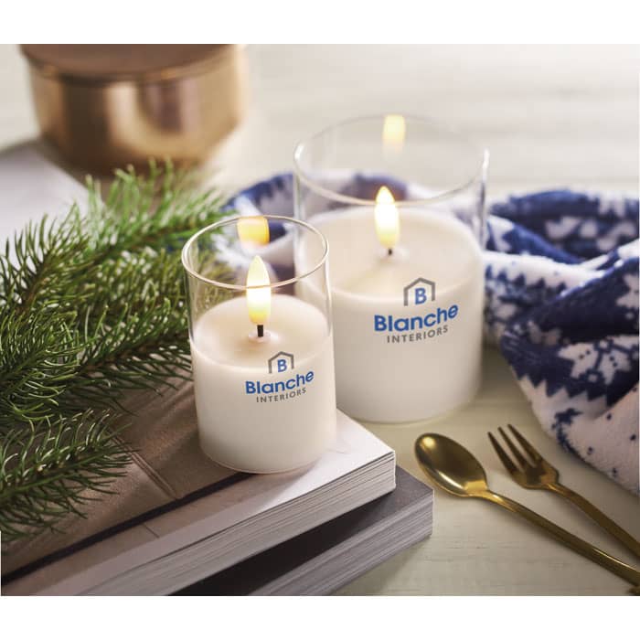 Two candles in clear glass holders, one larger and one smaller, with a lighted flame, set on a table with greenery.