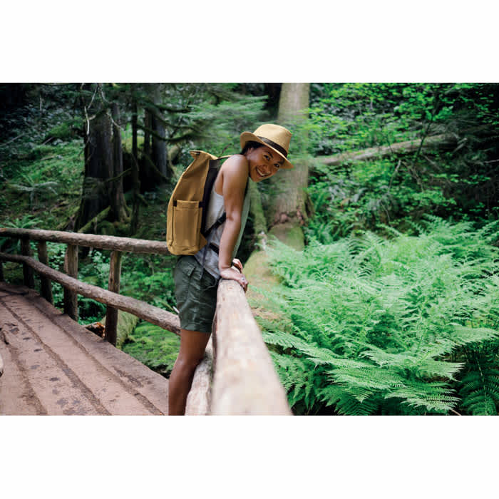A person wearing a hat and shorts leans on a wooden railing in a lush green forest, with ferns in the background.