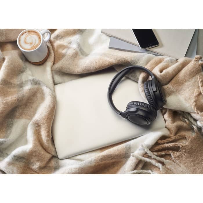 A flat lay featuring a laptop, black headphones, a coffee cup, and a patterned blanket in neutral colours.