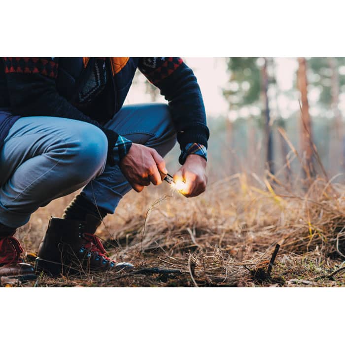 A person kneeling on the ground, using a metal tool to create a spark in a forested area.