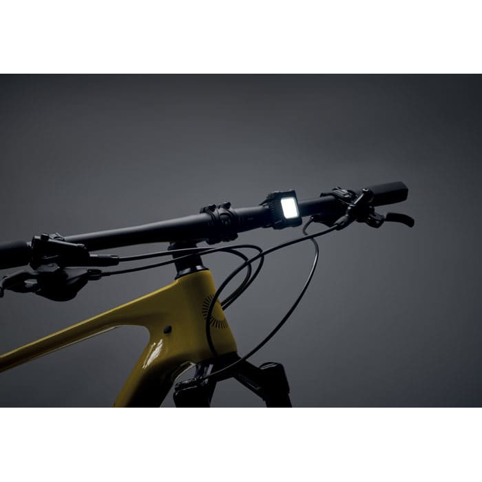 A bike handlebar with a yellow frame and a square light mounted on it, against a dark background.
