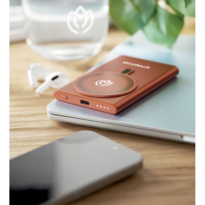 A portable charger in a copper colour, featuring a circular design, placed on a desk next to a phone and earbuds.