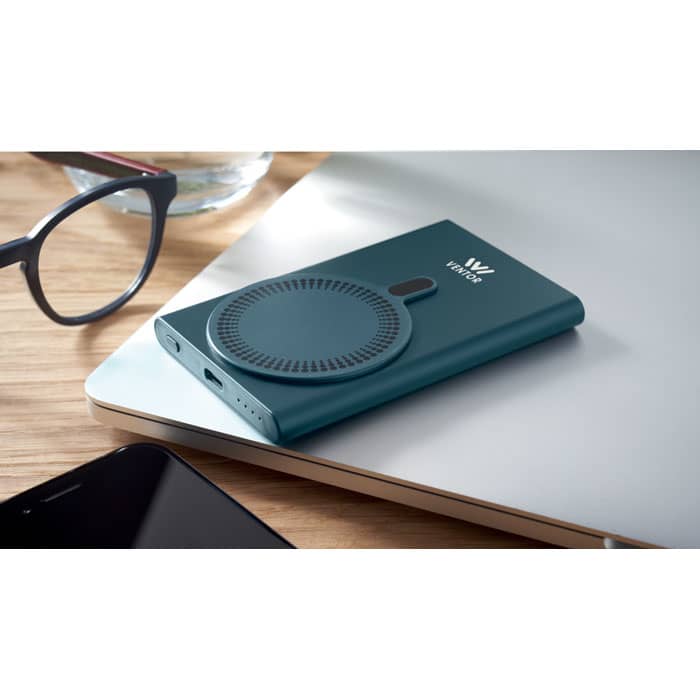 A teal portable charger with a circular design, placed on a wooden desk next to glasses and a laptop.