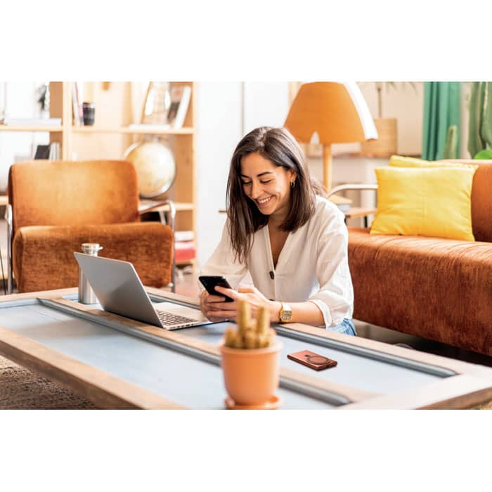 A woman with brown hair sits on a floor, using a phone and laptop beside a small potted plant on a light table.