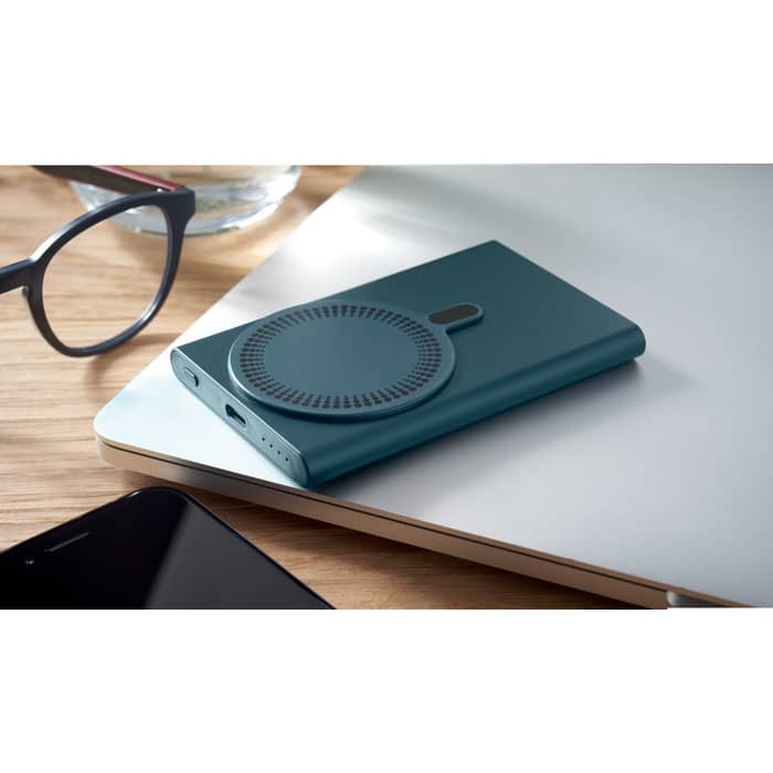 A teal drink bottle sits on a wooden table next to glasses and a laptop, featuring a circular design on one side.