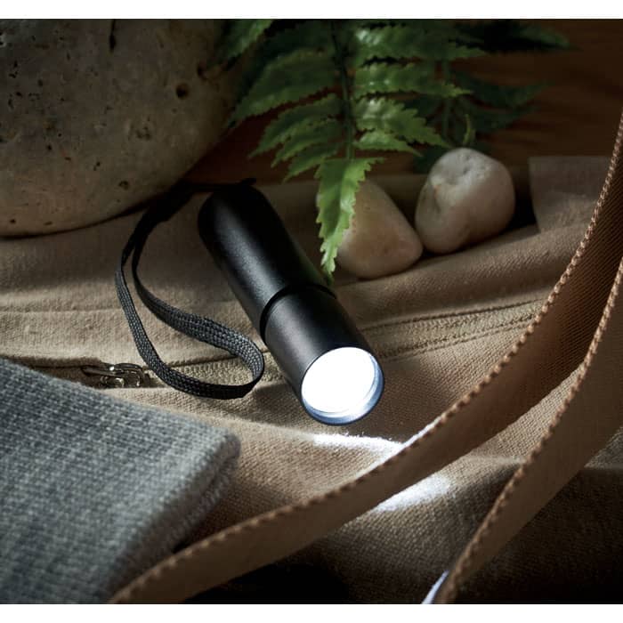 A black flashlight rests on a textured surface alongside pebbles and greenery. It features a strap for easy carrying.