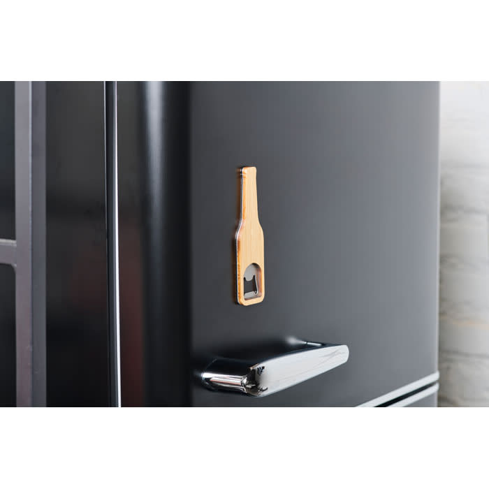 A bottle opener shaped like a drink bottle attached to a black fridge, made from wood. It has a round cutout and a simple design.