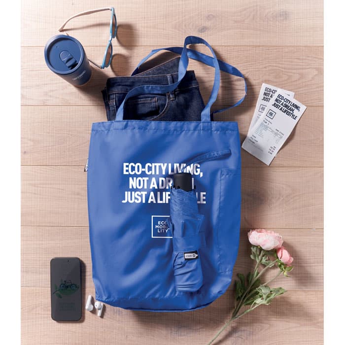 A blue tote bag with sunglasses, a drink tumbler, jeans, tickets, a phone, and a flower. The bag has a logo.