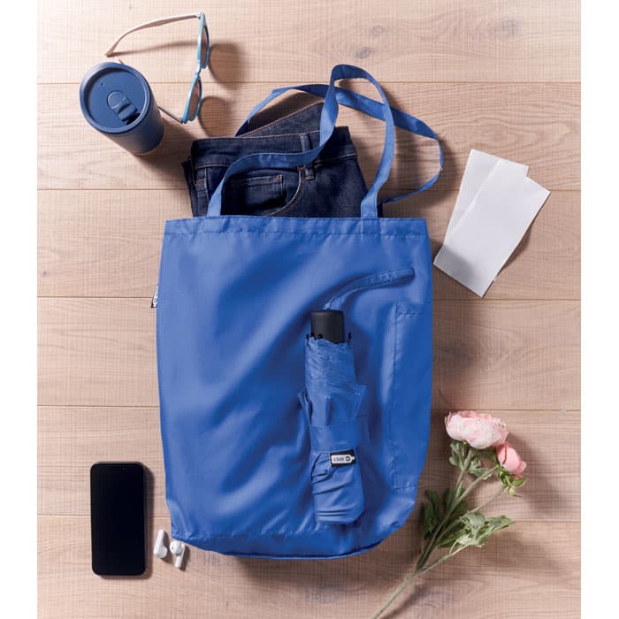 A blue tote bag with a logo, containing sunglasses, jeans, a drink bottle, a smartphone, and flowers.