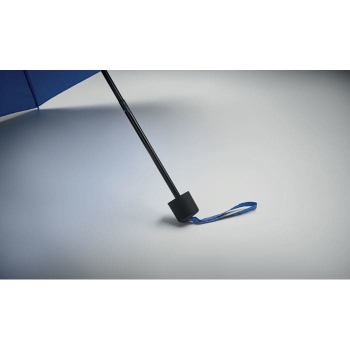A blue umbrella with a black handle and a strap, resting on a smooth, light surface. It features a logo.