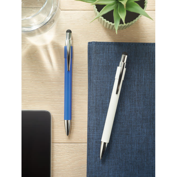 Two pens, one blue and one white, are placed on a wooden surface beside a notebook and a glass of water.