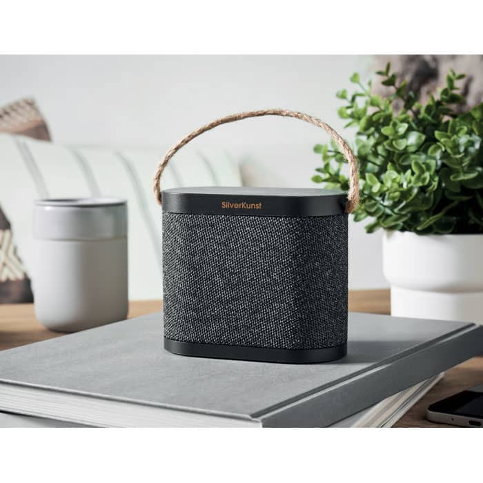 A portable speaker with a grey fabric exterior and a rope handle, resting on a stack of books beside a plant.