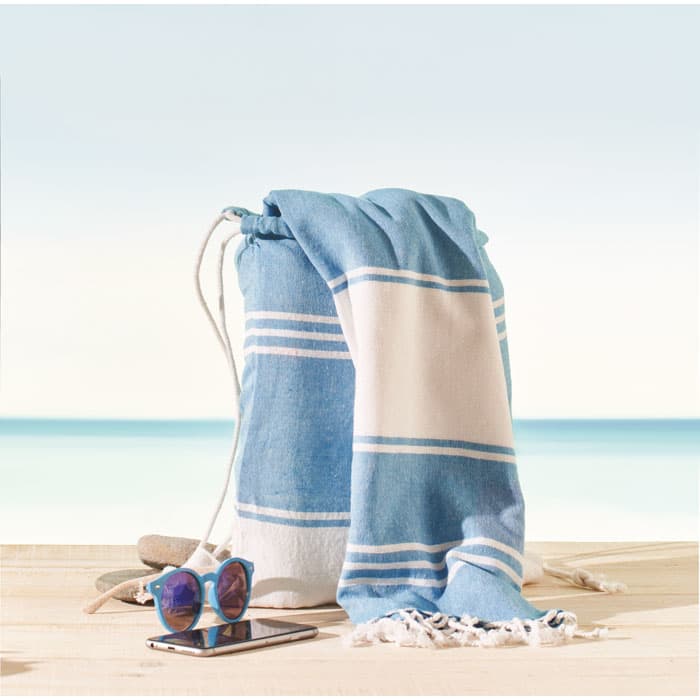 A beach bag with a blue and white towel, sunglasses, and a smartphone, set against a sandy shore and ocean background.