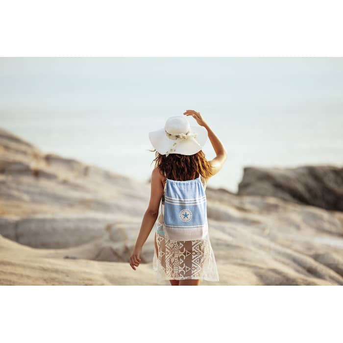A person wearing a wide-brimmed hat carries a blue and white drawstring backpack while walking along a rocky shore.