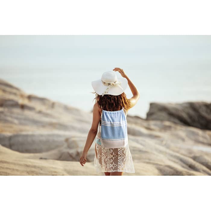 A woman with a white hat and a blue backpack walks along rocky terrain by the water. The background features a serene ocean view.