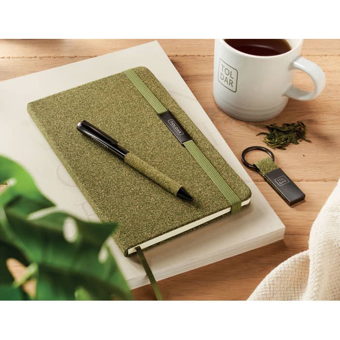 A green notebook with a pen and keychain, accompanied by a coffee cup, laid on a wooden surface with plants.