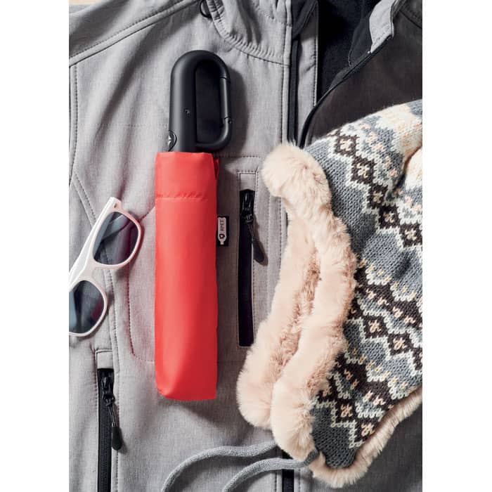 A red umbrella case alongside sunglasses and a textured scarf, resting on a grey jacket. The scene includes a logo.