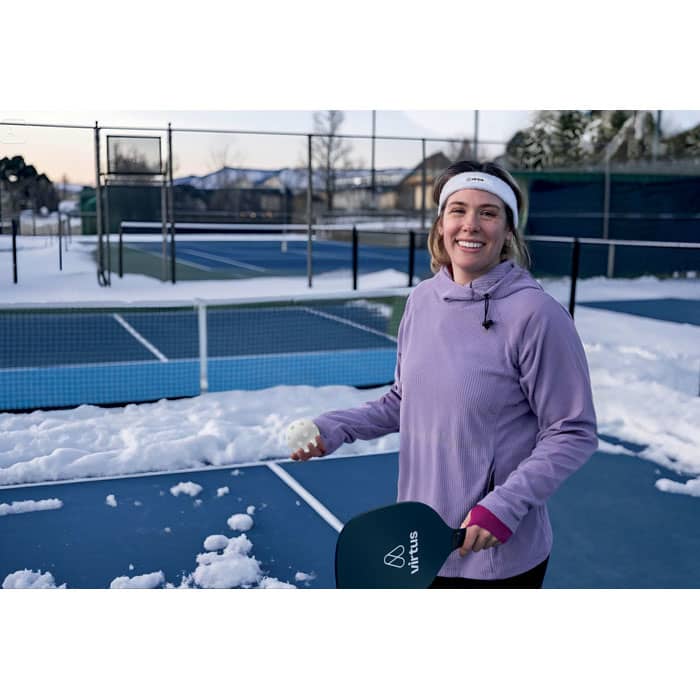 A smiling person in a purple hoodie holds a paddle on a snowy tennis court, with snow-covered courts in the background.