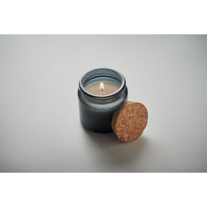 A candle in a dark glass jar, featuring a cork lid, with a lit wick illuminating the interior.