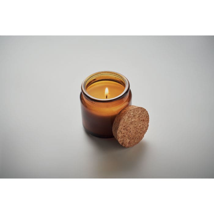 A candle in an amber glass jar with a cork lid, featuring a lit wick and warm, flickering flame.