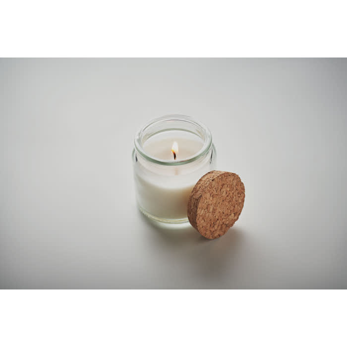 A glass jar candle with a lit wick and a cork lid placed beside it. The candle is white.