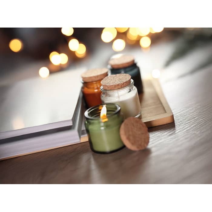 A display of four small candles in glass jars with cork lids, in green, white, orange, and dark blue, on a wooden tray.