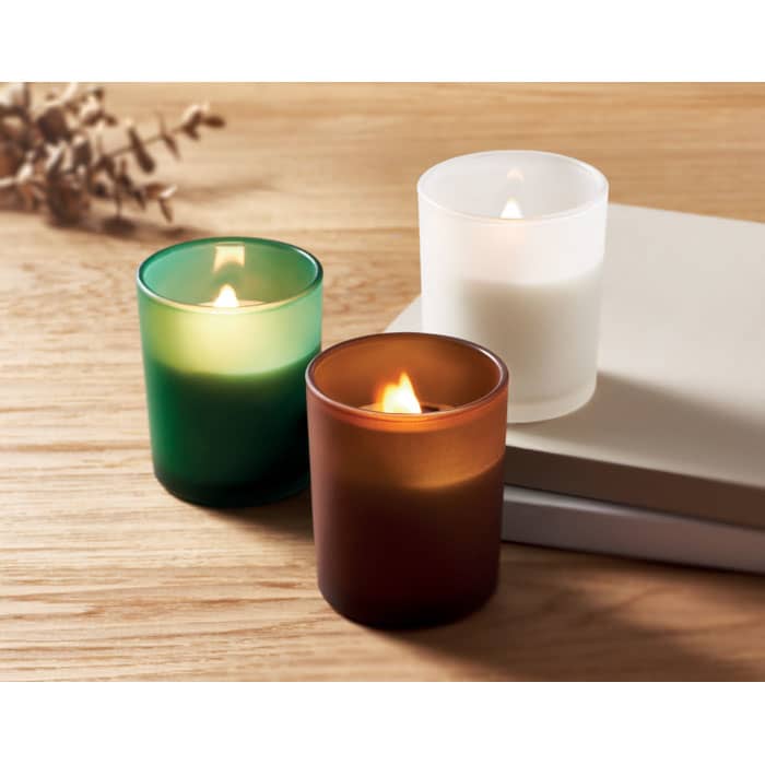 Three scented candles in green, brown, and white glass holders, arranged on a wooden surface with a few books.