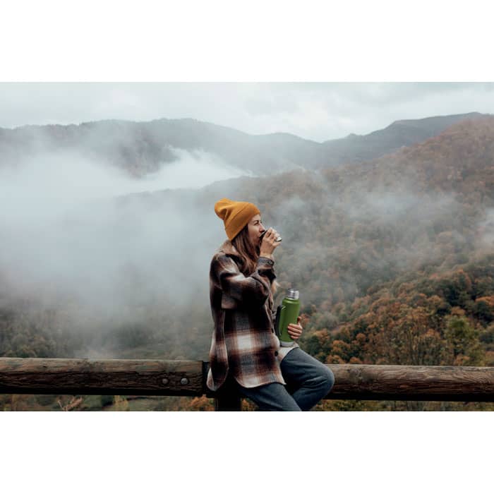 A drink bottle in green held by a woman wearing a yellow beanie, sitting against a wooden rail with misty mountains in the background.
