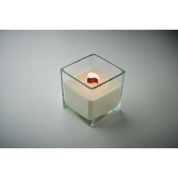 A square glass candle holder with a white wax candle burning inside. The flame is flickering gently.