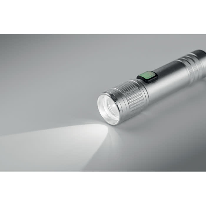 A silver torch with a textured grip and a green button, emitting a bright beam of light.