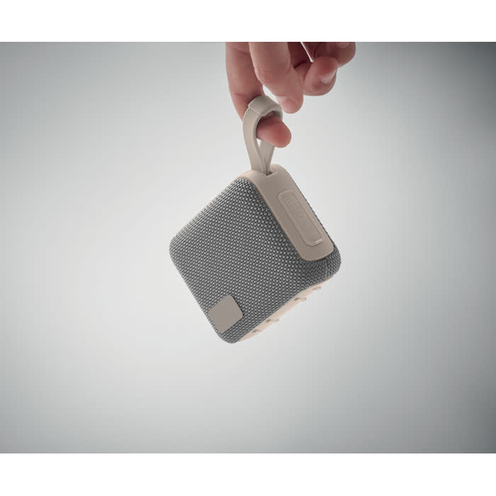 A portable speaker in grey fabric with a handle, being held by a hand against a light background.