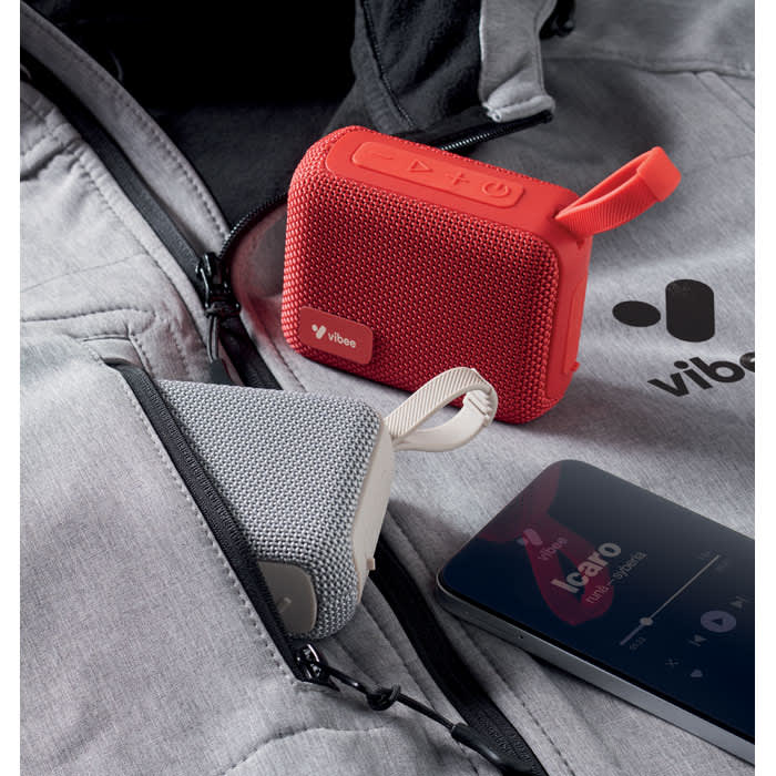 A compact red speaker and a grey speaker are placed next to a smartphone, all set against a grey jacket background.